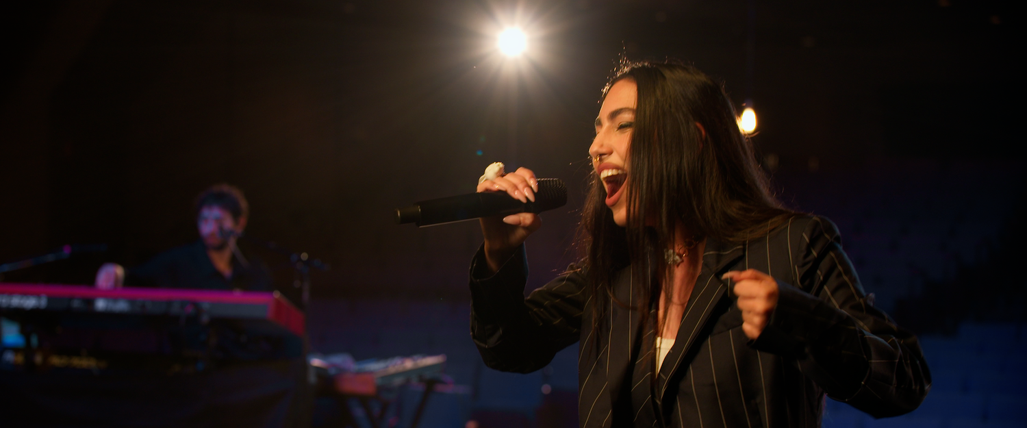 Maria Jose Llergo featured image. María José Llergo sings on stage in a dark pinstripe suit, with a keyboardist in the background.
