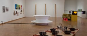 Contemporary art gallery with various installations, including a large white bowl sculpture, wall-mounted artworks, a digital clock, and a table set with dishes.