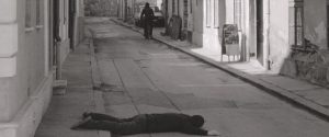 A person lies face down on a deserted street while another person approaches on a bicycle in the background.