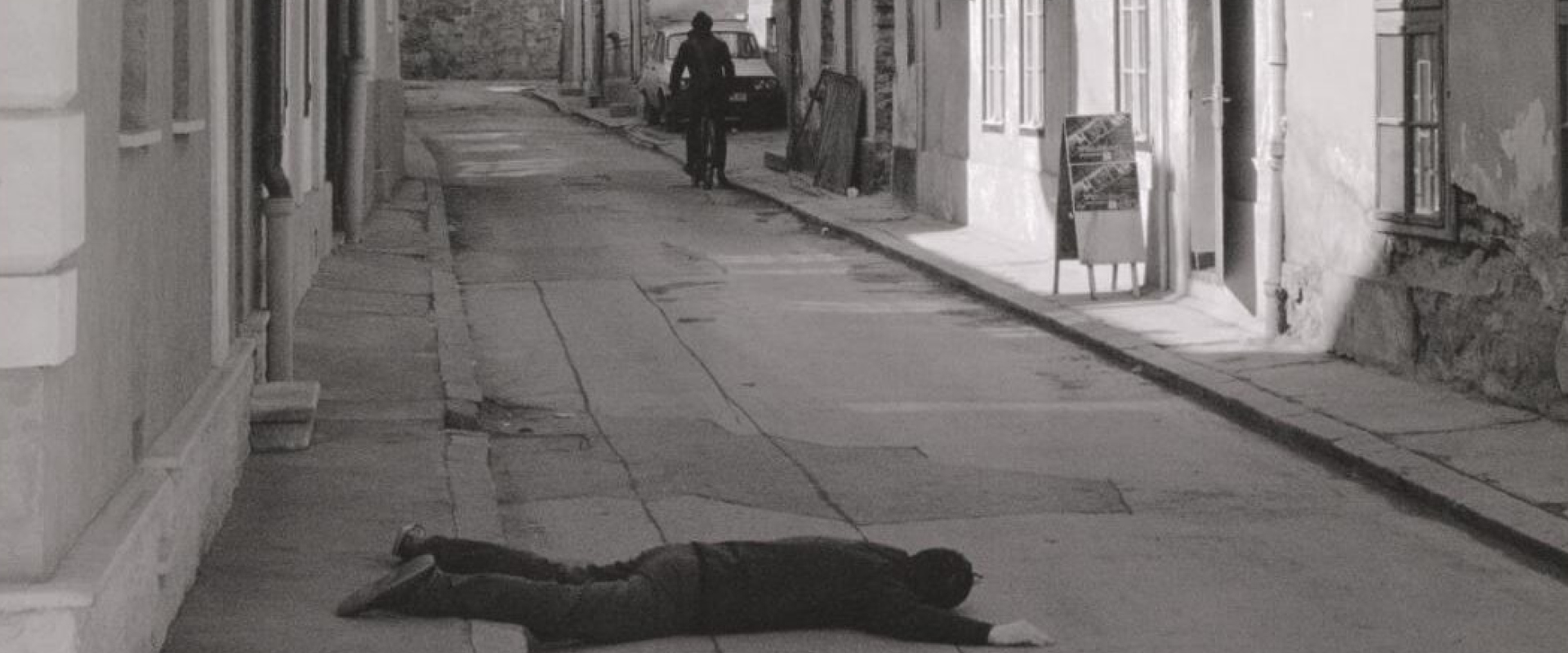 A person lies face down on a deserted street while another person approaches on a bicycle in the background.