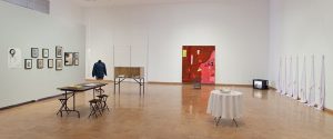 Art gallery with framed artworks on the wall, a denim jacket display, tables with objects, a red abstract painting, white draped items, and a small TV on the floor.