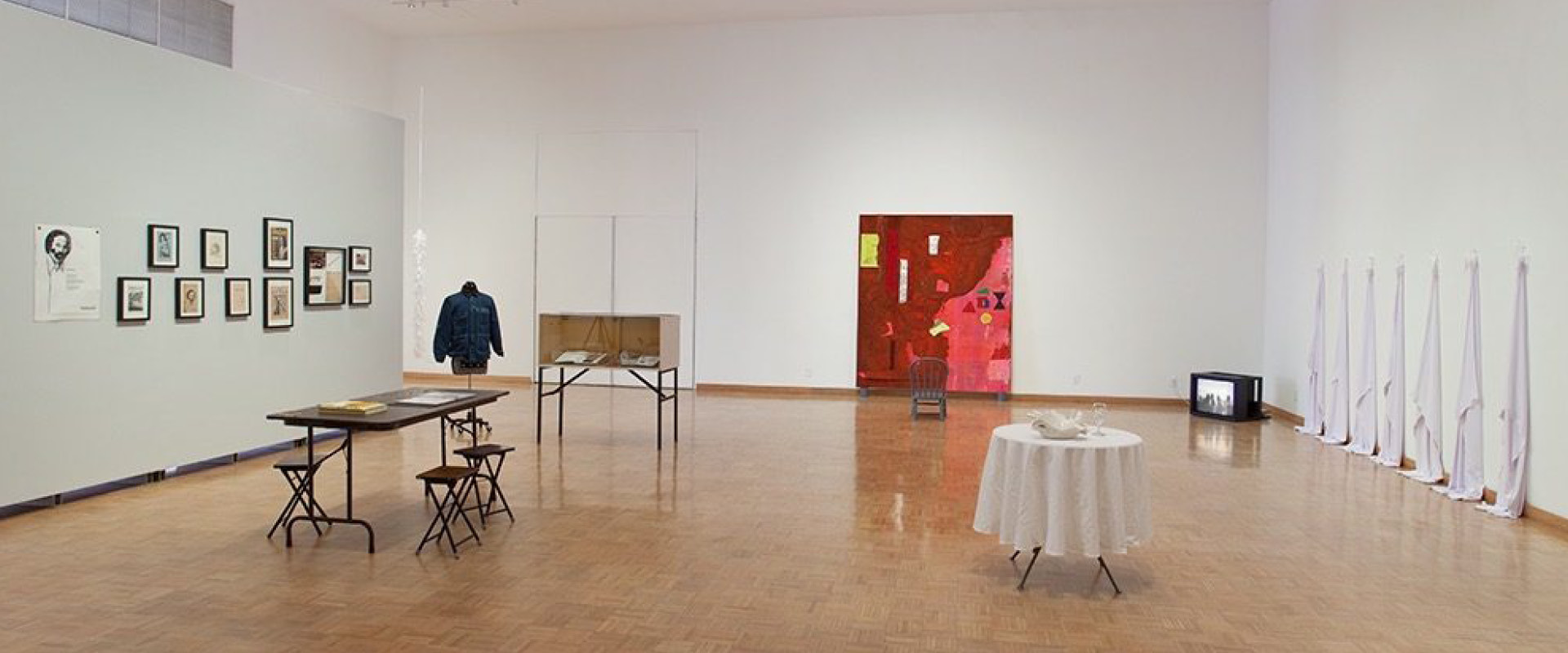 Art gallery with framed artworks on the wall, a denim jacket display, tables with objects, a red abstract painting, white draped items, and a small TV on the floor.