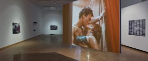 An art gallery with a large projected film scene of a couple kissing in a shower, and several framed artworks on the surrounding white walls.