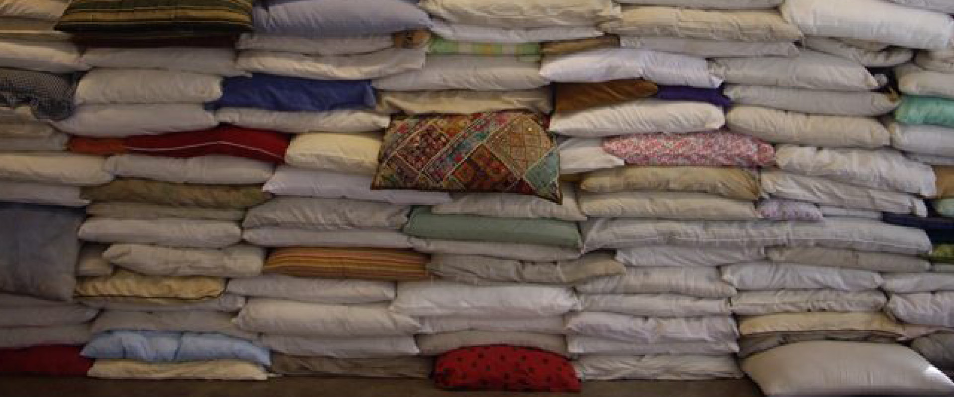 A large stack of variously colored and patterned pillows and cushions piled against a wall in several uneven rows.