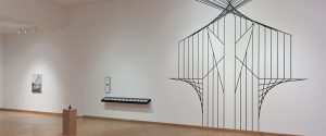 A minimalist art gallery with a large geometric wall mural, framed artworks, and a small sculpture on a pedestal displayed in a spacious, well-lit room.