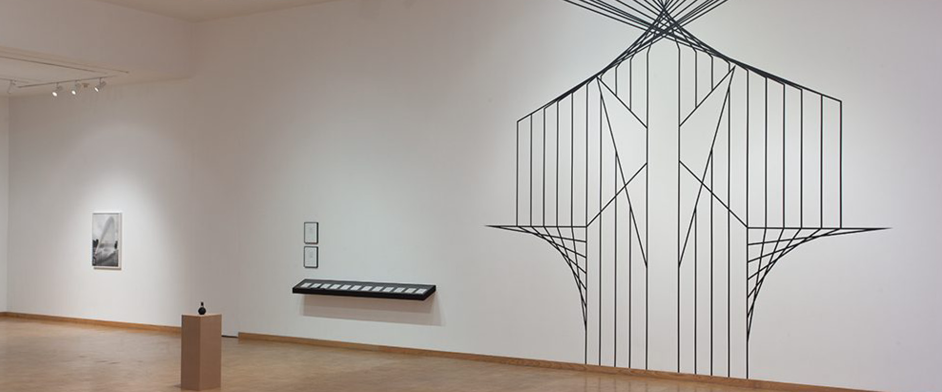 A minimalist art gallery with a large geometric wall mural, framed artworks, and a small sculpture on a pedestal displayed in a spacious, well-lit room.