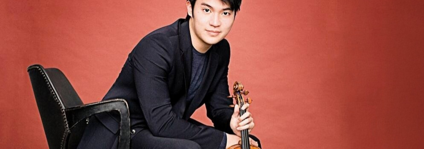 Violinist Ray Chen sits holding his violin and gazes at the camera against a warm red background.