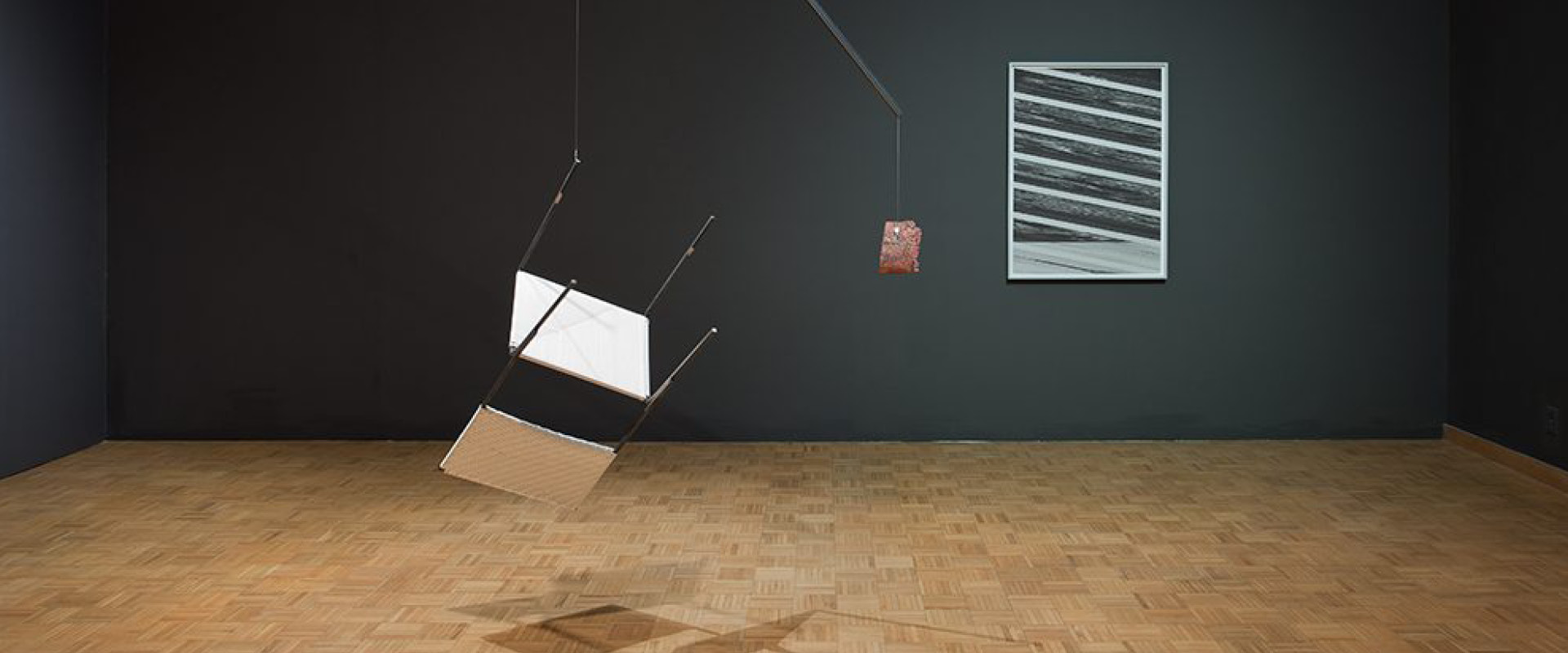 A mobile sculpture with suspended geometric shapes hangs in a gallery space with parquet flooring; a striped abstract painting is displayed on a dark wall.