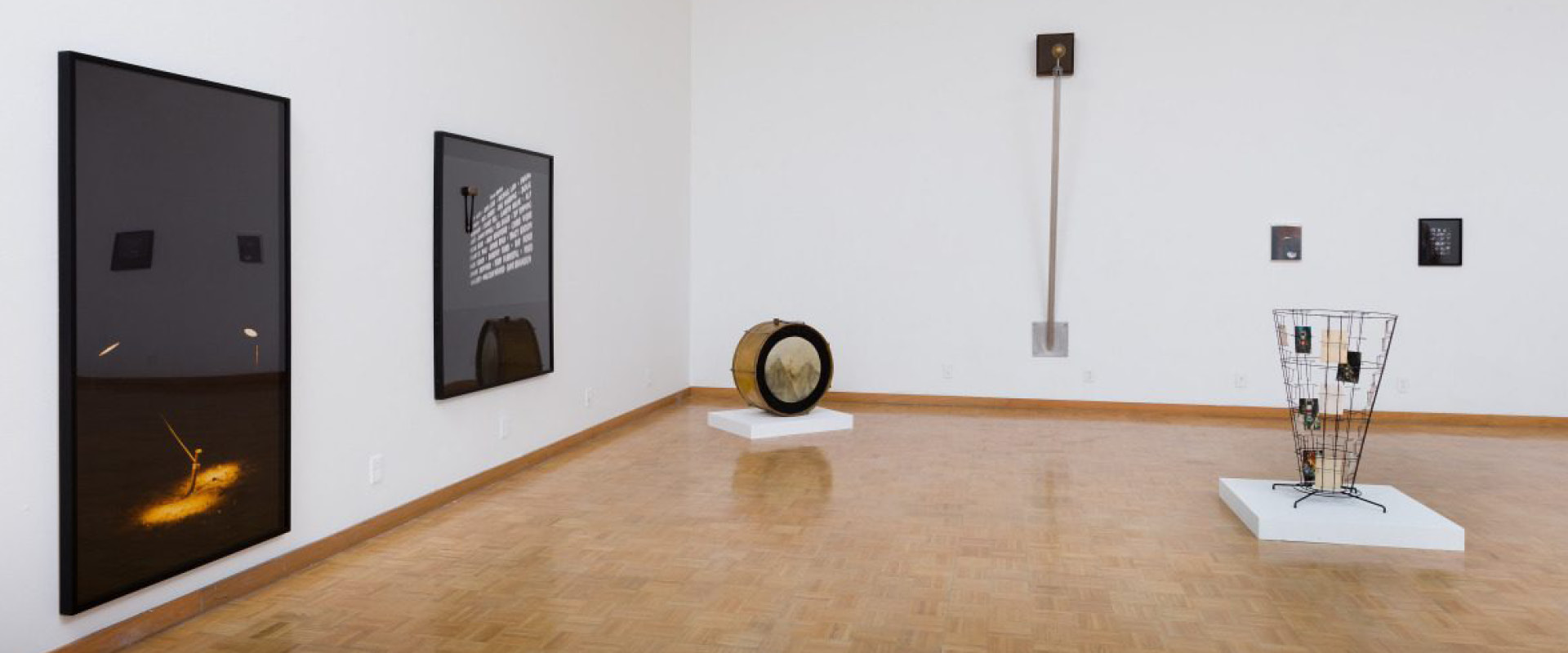 Gallery space with contemporary art pieces displayed on white walls and floor, including framed images, a large drum, a tall sculpture, and a wire basket on a pedestal.