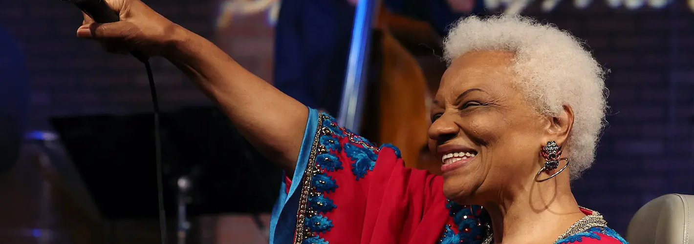 Jazz vocalist Barbara Morrison smiles broadly and raises a microphone overhead while performing in a vibrant red-and-blue embroidered dress.