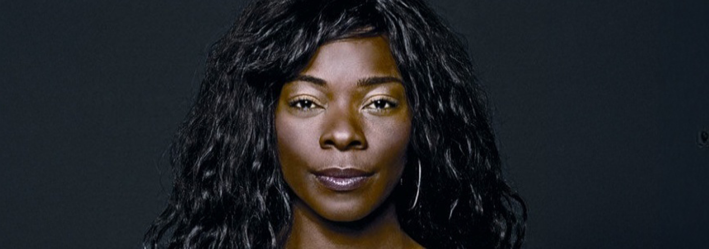 Close-up portrait of singer Buika looking directly at the camera, her long, wavy black hair contrasting with a dark background.