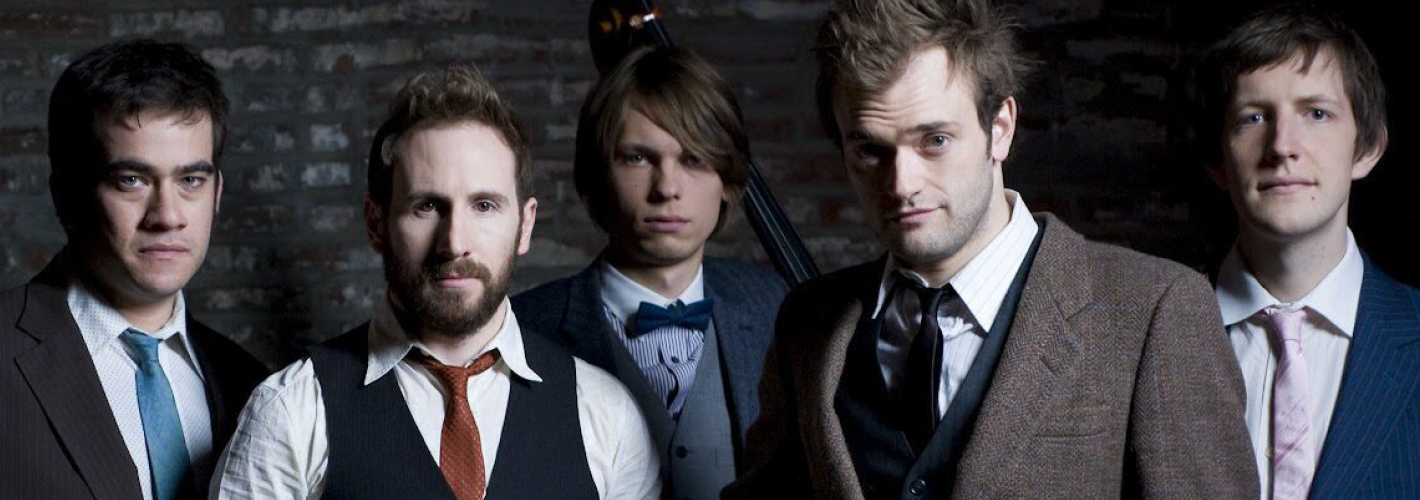 The five members of bluegrass band Punch Brothers pose in suits with string instruments against a dark brick wall.
