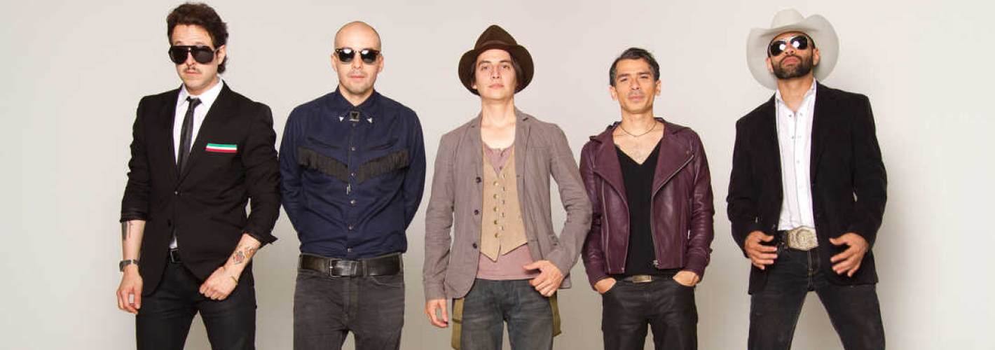 The five members of Mexican rock band Kinky pose in suits, jackets, hats, and sunglasses against a plain white background.