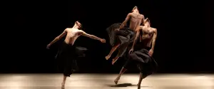 Three shirtless dancers wearing flowing black skirts perform expressive, dynamic movements on a dimly lit stage against a dark background.