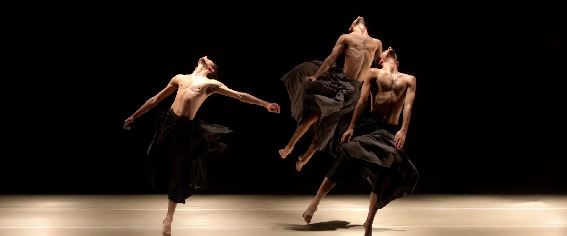 Three shirtless dancers wearing flowing black skirts perform expressive, dynamic movements on a dimly lit stage against a dark background.