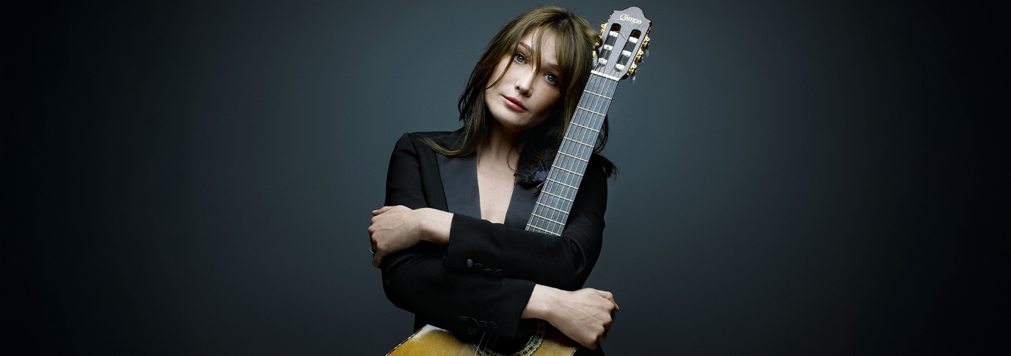 Singer-songwriter Carla Bruni embraces a classical guitar while gazing at the camera against a dark gray background.