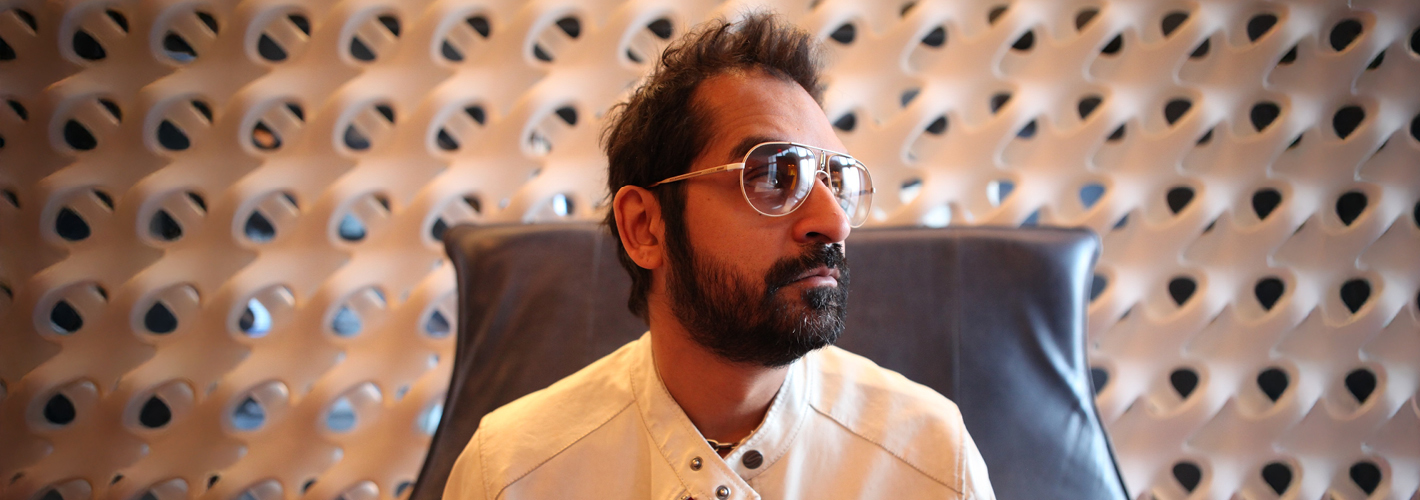 Karsh Kale wearing sunglasses and a light-colored jacket, seated against a modern geometric-patterned backdrop.