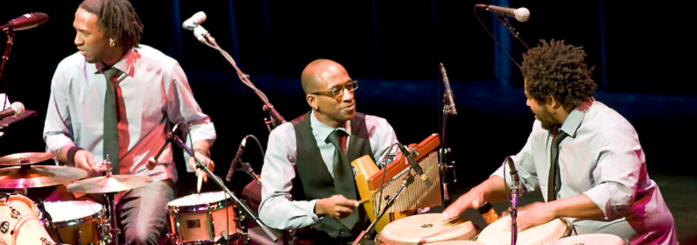 Three percussionists perform together onstage, playing a drum kit, chimes, and hand drums beneath microphones.