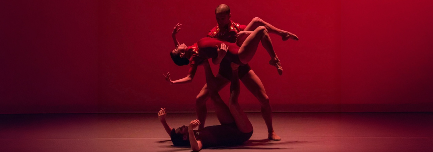 Four dancers form an intertwined sculptural pose onstage, dramatically illuminated by deep red lighting.