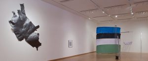 A contemporary art gallery with a large black abstract piece on the left wall, a framed artwork, and colorful stacked cylindrical sculptures in the background.