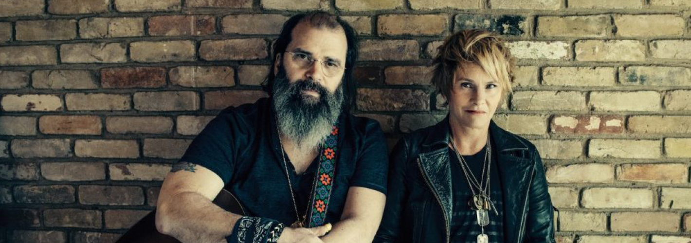 Steve Earle with a beard a guitar sitting alongside with Shawn Colvin against a rustic brick wall, both exuding a cool, artistic vibe.