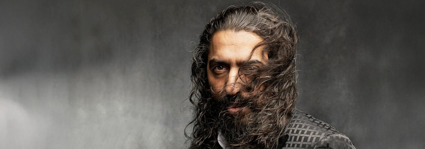 Diego el Cigala with long curly hair and beard against a textured gray background, exuding confidence and intensity.