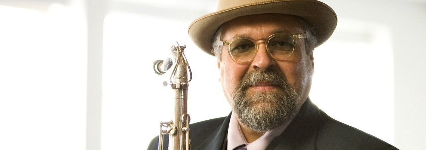 Joe Lovano with a beard and glasses, wearing a fedora hat, a tie, and a suit, holding a saxophone and smiling confidently.