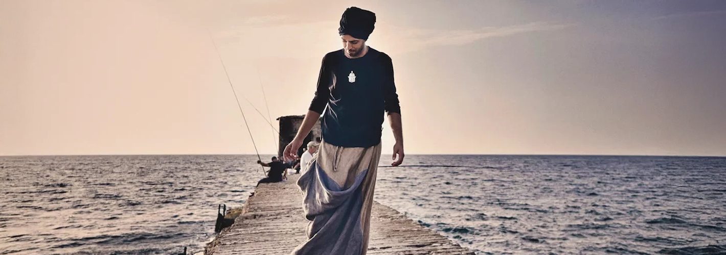 Musician Idan Raichel walks along a stone pier at sunset, surrounded by the sea as people fish in the distance.