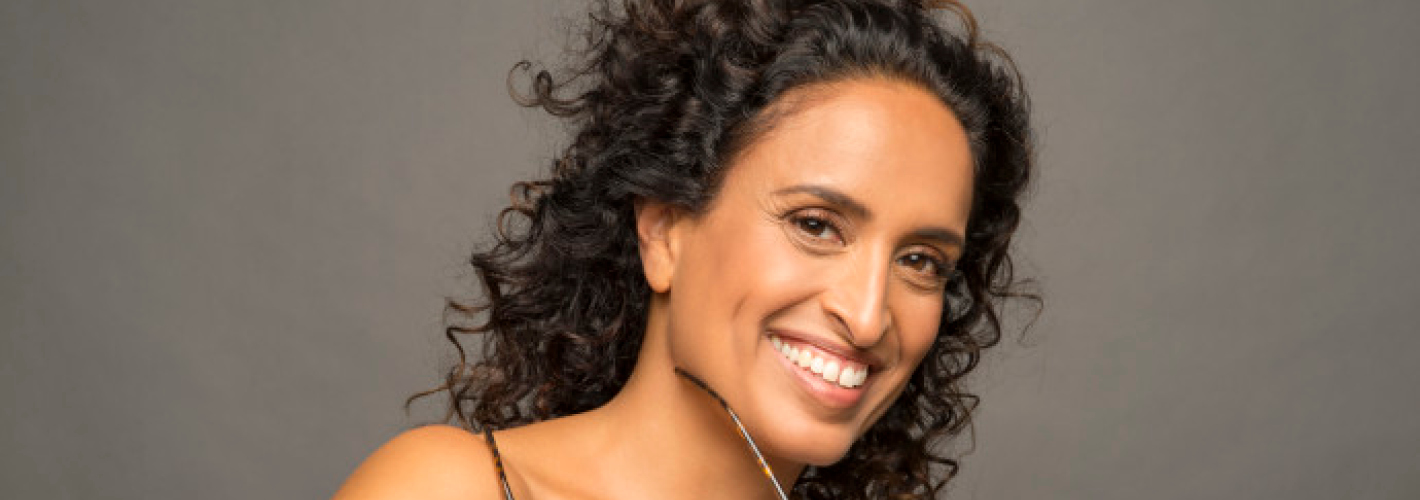 Portrait of a smiling woman, Achinoam NiNi, with curly dark hair holding eyeglasses near her chin against a gray background.