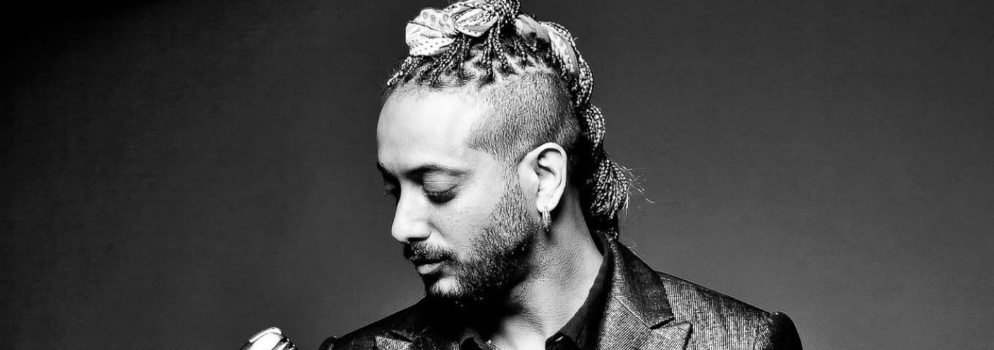 Monochrome image of a stylish man holding a microphone, featuring a braided hairstyle and a unique head accessory, set against a dark background, exuding an artistic and edgy vibe.