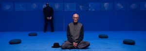 Ying Ming Tu in a dark tunic sitting cross-legged on a vibrant blue carpeted floor with a hat placed beside him, while another person stands in the background looking at framed portraits on the wall.