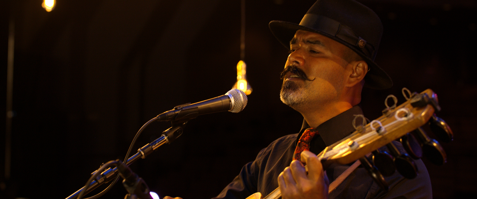 A musician with a mustache and beard, wearing a black hat and shirt, passionately sings and plays a guitar on stage. Warm lighting highlights him against a dark background, with an exposed bulb hanging nearby.