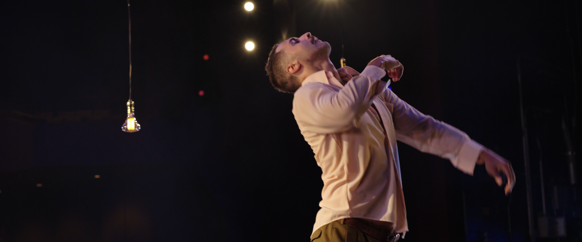 A person in a light shirt dances expressively on stage under a hanging light bulb. The background is dark, with a few lights creating a dramatic, moody atmosphere.