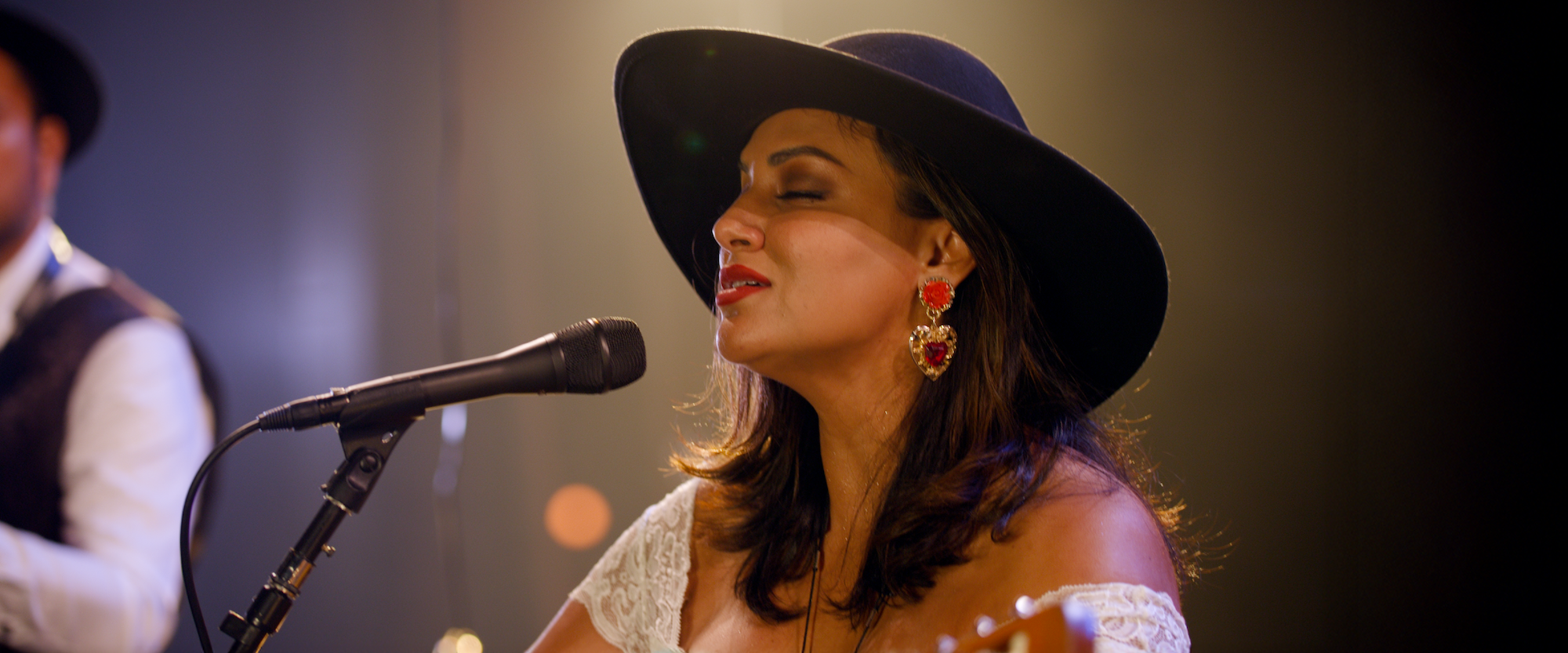 A woman wearing a black hat and large red earrings sings passionately into a microphone on stage. She has long dark hair and is illuminated by warm stage lights. A blurred musician is in the background.