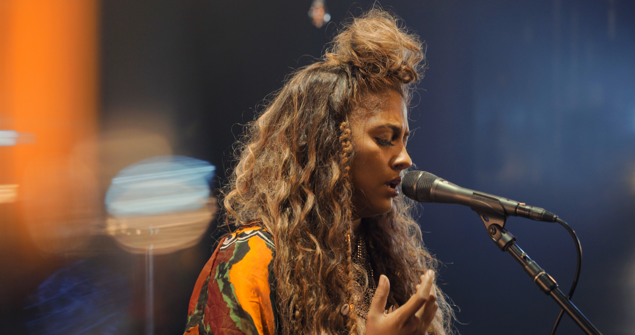 Candice Pillay sings passionately on stage in a colorful outfit, with blurred lights behind her.