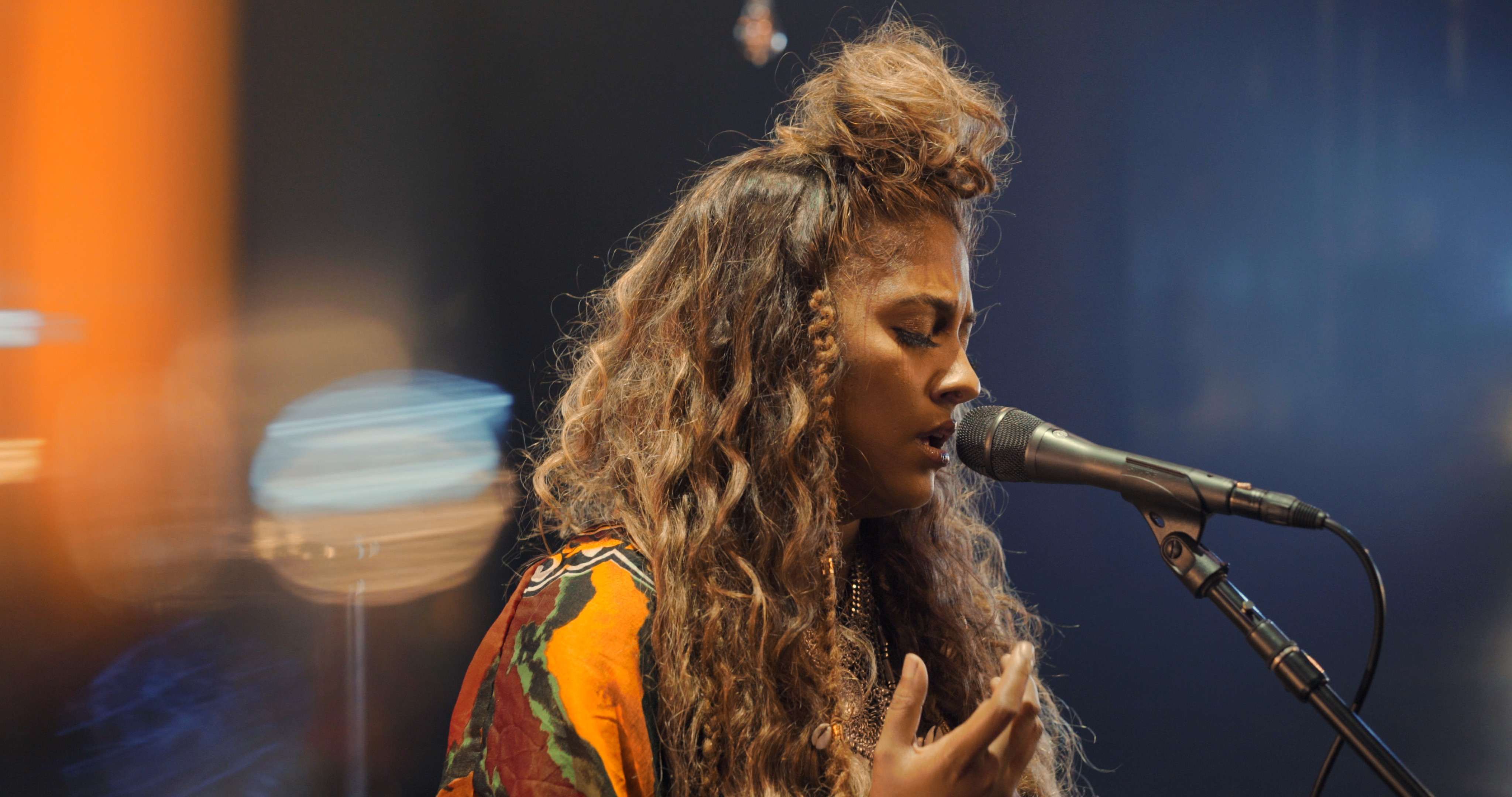 A woman with long, curly hair sings passionately into a microphone on stage. She wears a colorful outfit and appears deeply immersed in her performance, with blurred lights in the background creating an atmospheric effect.