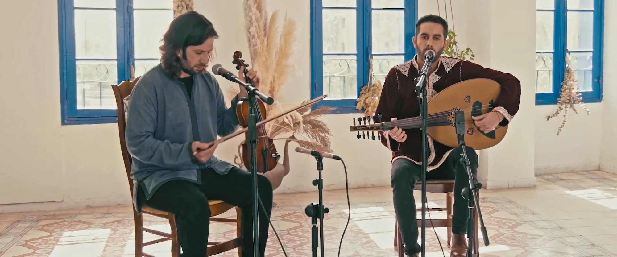 Two musicians perform indoors, framed by large windows. One plays a violin while looking at sheet music, and the other sings into a microphone while playing an oud. The setting is bright and decorated with dried plants.