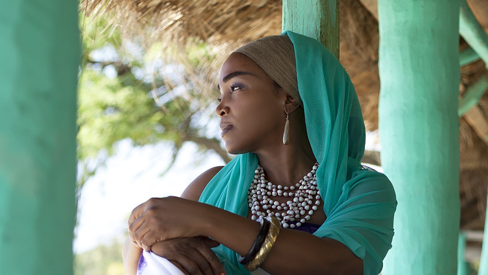 Emeline Michel in a teal headscarf and matching top, adorned with a bold white necklace, gazes into the distance, surrounded by the serene setting of a thatched structure.