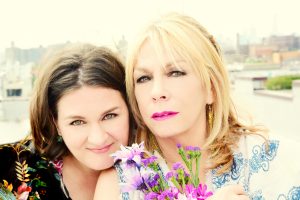 Madeleine Peyroux and Rickie Lee Jones sharing a close moment, with one holding a bouquet of flowers, set against an urban rooftop backdrop.