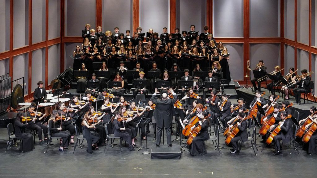 An orchestra in full performance, led by a conductor with a chorus in the background.