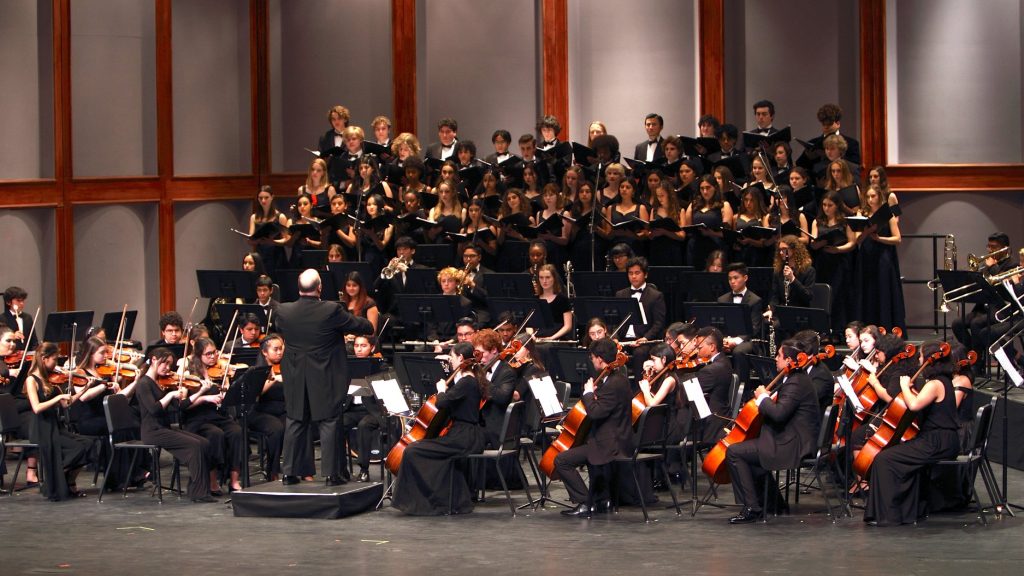 A symphony orchestra and choir perform onstage, with musicians playing various instruments and a conductor leading the ensemble.