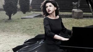 A woman in an elegant black dress playing a grand piano outdoors, with trees and a staircase in the background, creating a serene and artistic atmosphere.