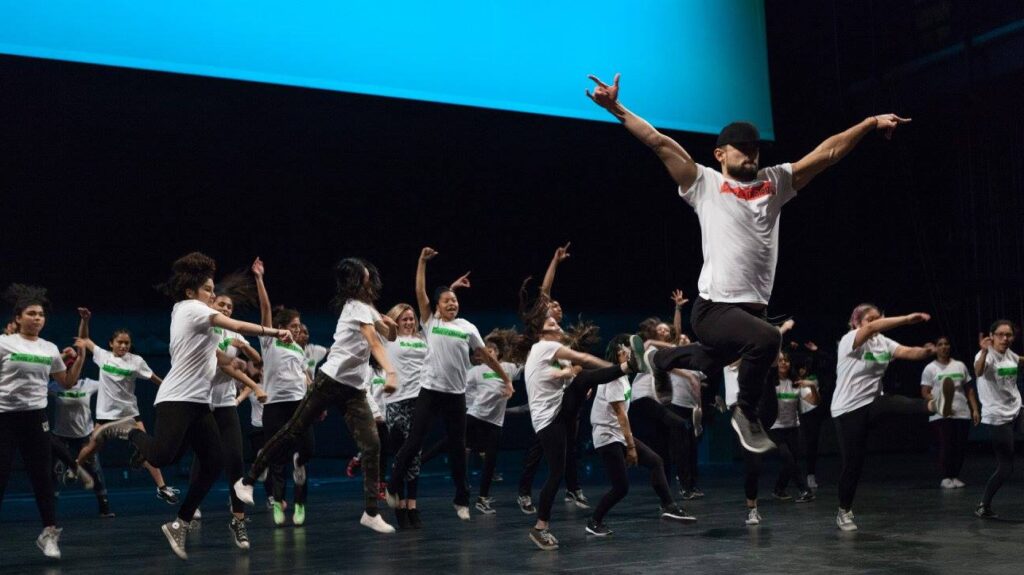 A dynamic group of dancers rehearsing on stage, following the lead of a central figure who commands attention with arms outstretched and a powerful stance.