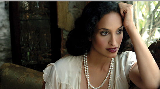 A pensive woman with dark hair and classic makeup, wearing a white blouse and a strand of pearls, gazes out the window in a room with rustic charm.