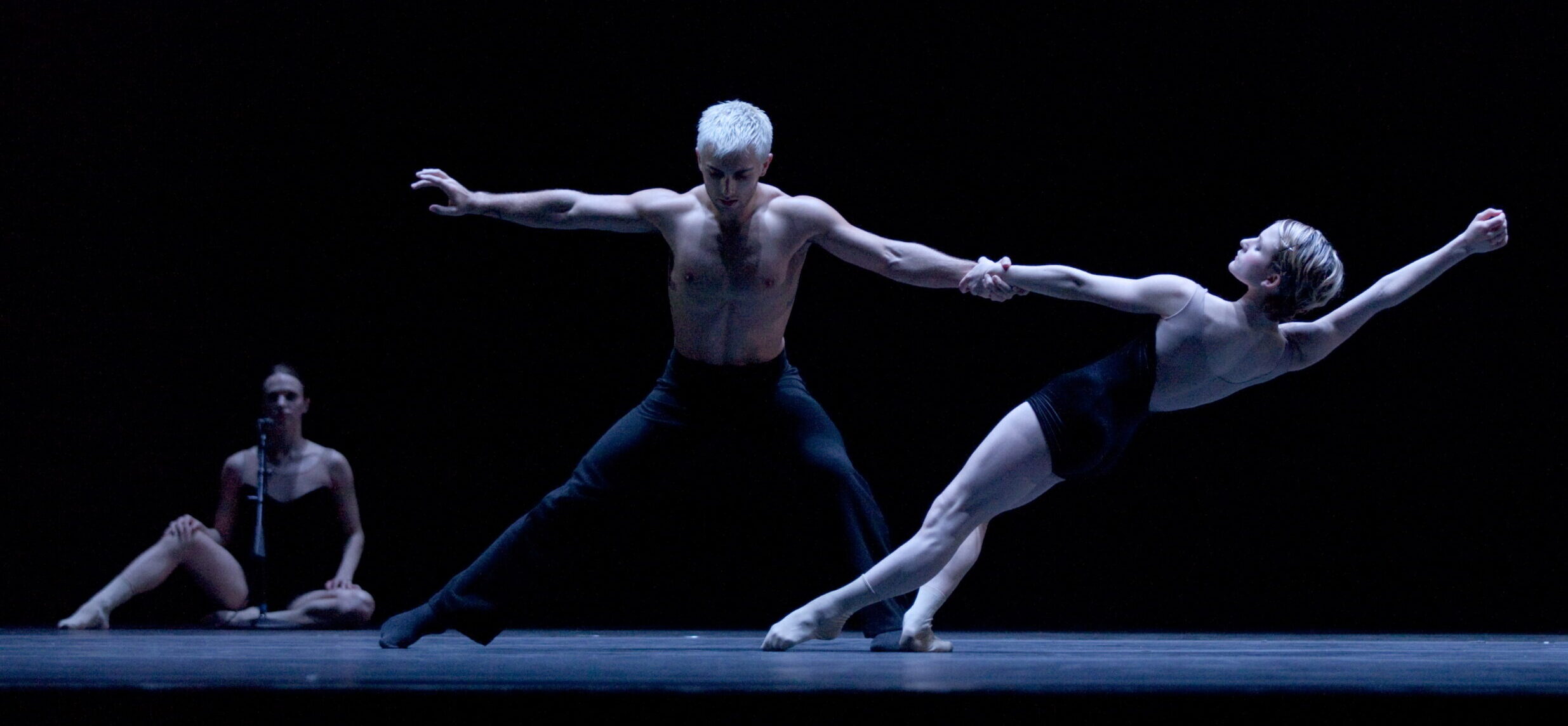A pair of ballet dancers perform a contemporary duet on stage, their movement capturing a moment of intense emotion and physical expression, while a solitary figure watches from the background.