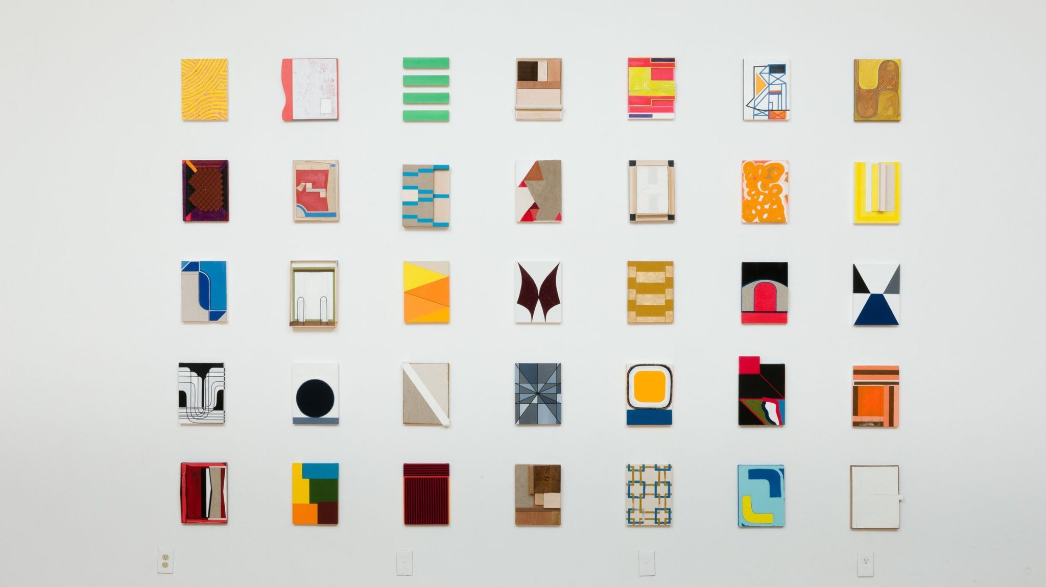 A symmetrical arrangement of various colorful abstract artworks on a white wall, providing an eclectic display of patterns, shapes, and designs.