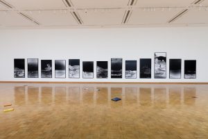 A spacious art gallery with a herringbone wood floor displays a series of large black and white photographic prints in linear arrangement on a clean white wall, with a scattering of colorful objects on the floor in the foreground.