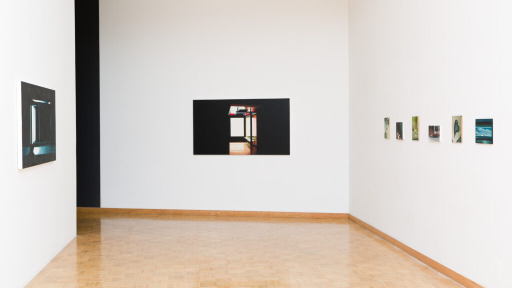 A minimalist art gallery interior with white walls featuring an assortment of framed photographs and paintings displayed at varying heights, with a parquet floor leading up to the artwork.