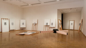 An art gallery room featuring a minimalistic exhibition with several framed artworks displayed on easels and two wooden benches for viewers to sit and contemplate the pieces.