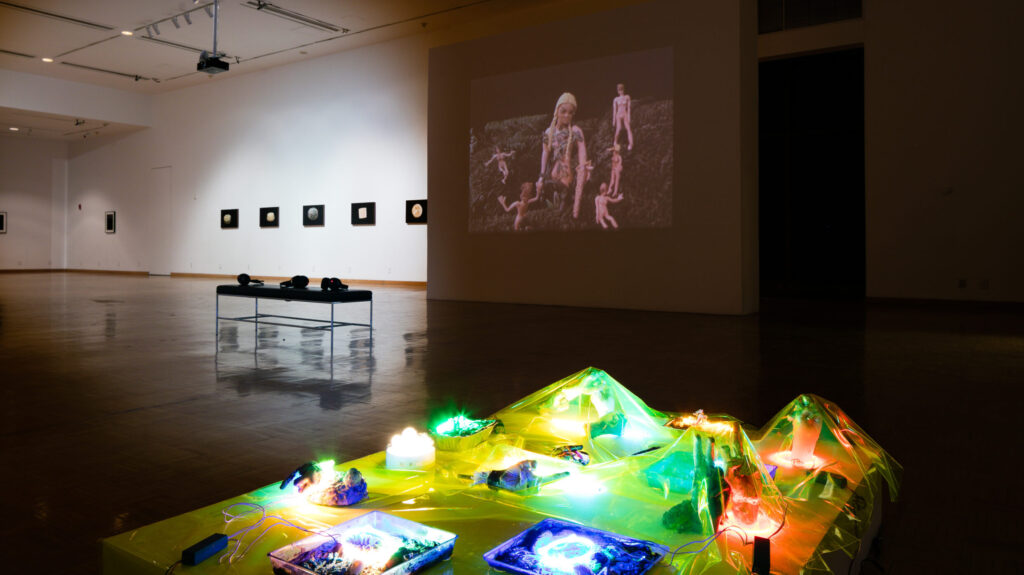 A contemporary art exhibition featuring a video projection and illuminated sculptural installations in a gallery setting, with framed artworks on the walls in the background.